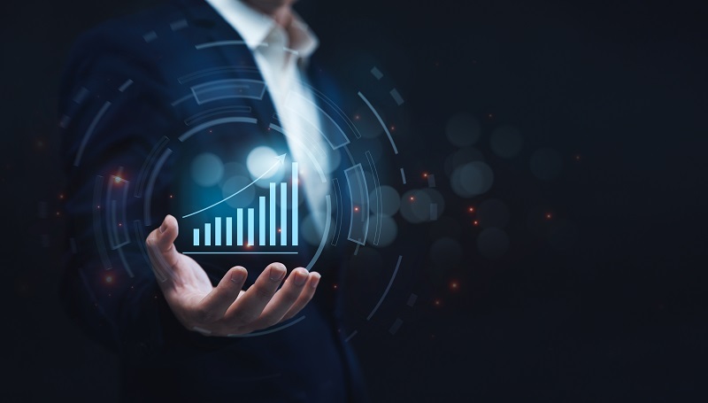 A businessman in a suit holds a glowing graphic of an upward-trending bar chart against a dark, blurred background.