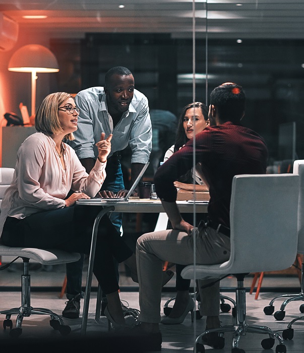 A diverse group of professionals engaged in discussion around a table, with a laptop, in a modern office setting.